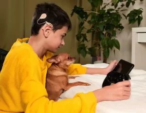 Teenage boy wearing a cochlear implant and looking at a tablet with a small dog